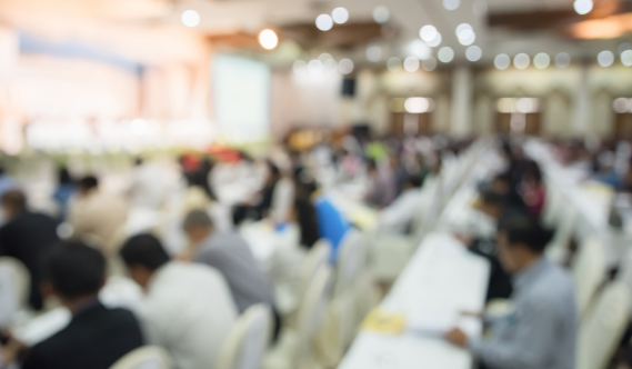 A blurred image of people seated at rows of tables in a large conference or seminar room.