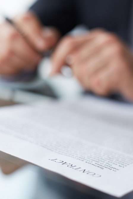 A person holding a pen is reviewing or signing a contract document on a desk. The word "CONTRACT" is clearly visible on the paper.