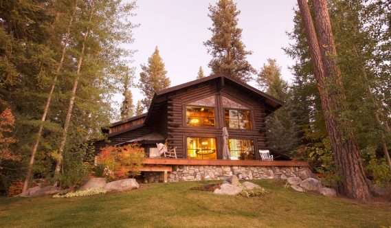 A two-story wooden log cabin with large windows and a stone foundation is surrounded by tall pine trees and a grassy yard at dusk.