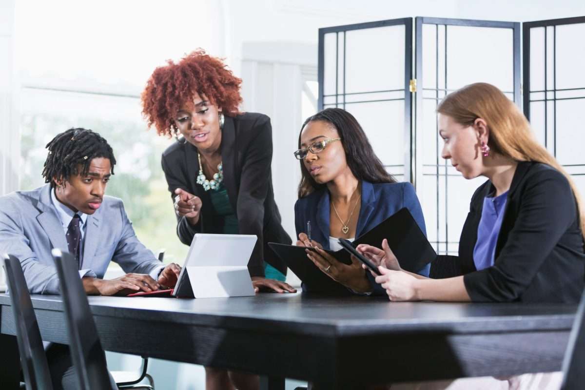 8 Four professionals sit at a table in an office, collaborating and reviewing documents and tablets during a meeting.