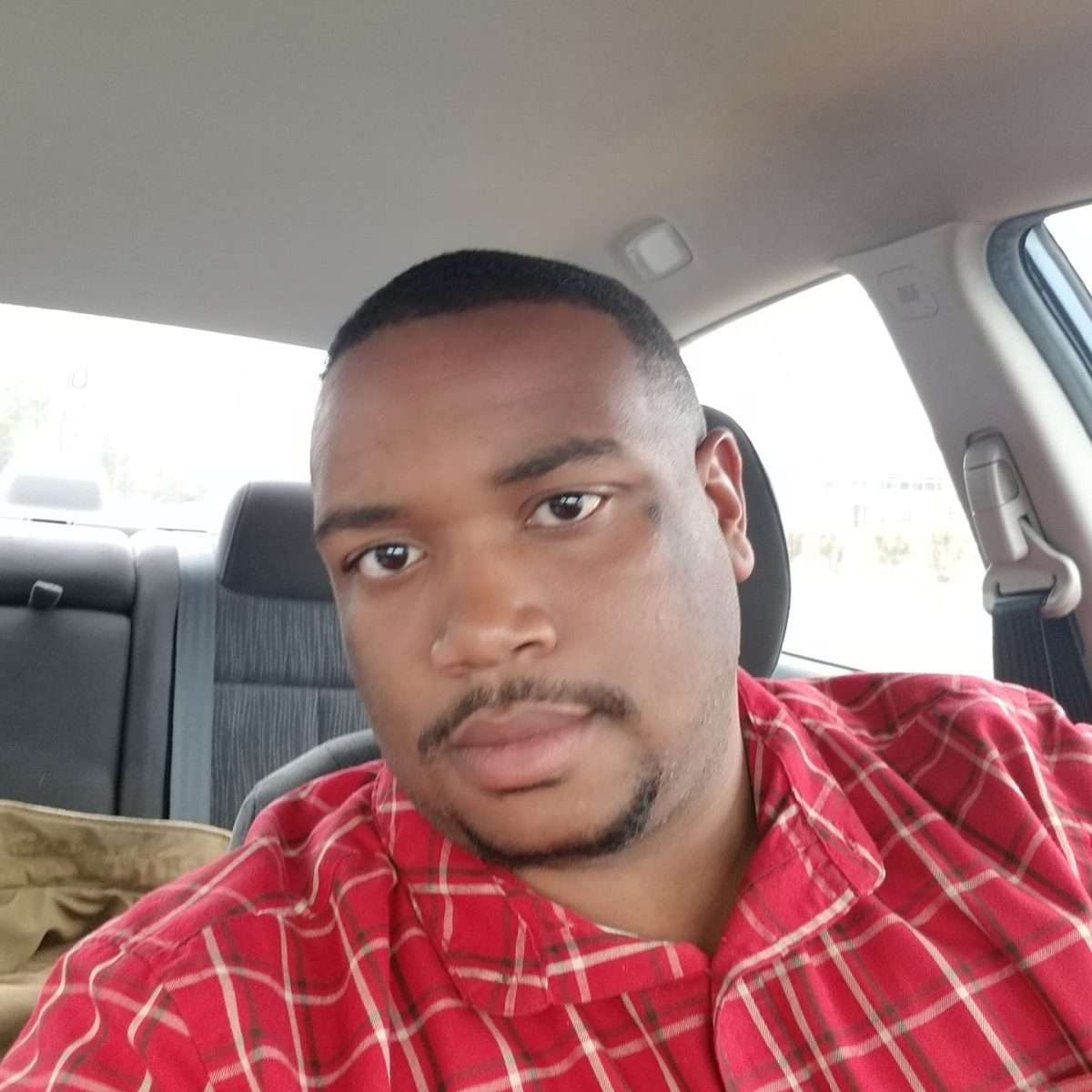 A man wearing a red plaid shirt sits in the driver's seat of a car, looking at the camera with a neutral expression.
