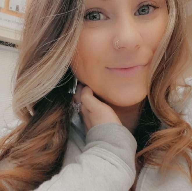 A woman with long, wavy brown hair and light highlights is smiling at the camera. She is wearing a gray top and a nose ring, with her hand resting near her neck.