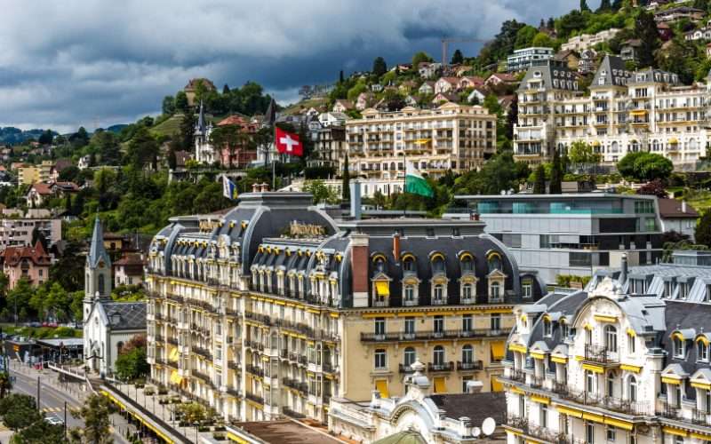 A grand historic hotel with a Swiss flag stands among hillside buildings and greenery under a cloudy sky.