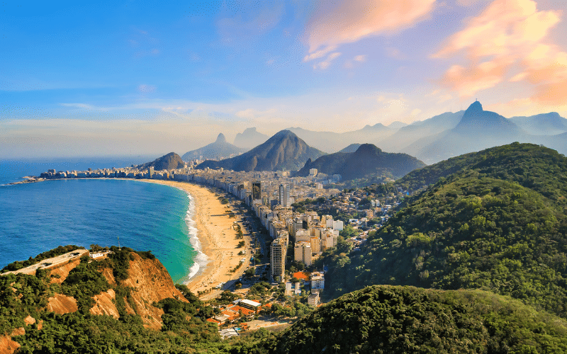 Aerial view of Rio de Janeiro showing a curving beach, city buildings, lush green hills, and mountains under a partly cloudy sky at sunset.