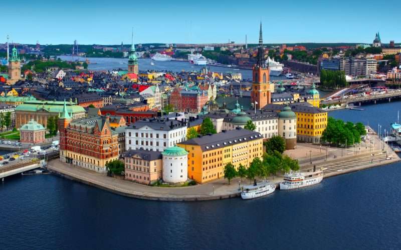 Aerial view of Stockholm&rsquo;s historic Old Town with colorful buildings, waterways, boats, and cityscape in the background under a clear blue sky.