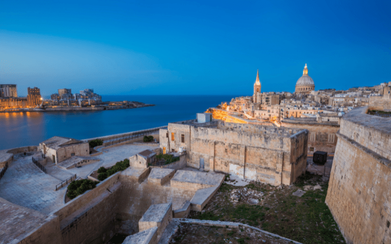 A view of Valletta, Malta at dusk, showing historic stone buildings, city lights, and a domed church near the waterfront.