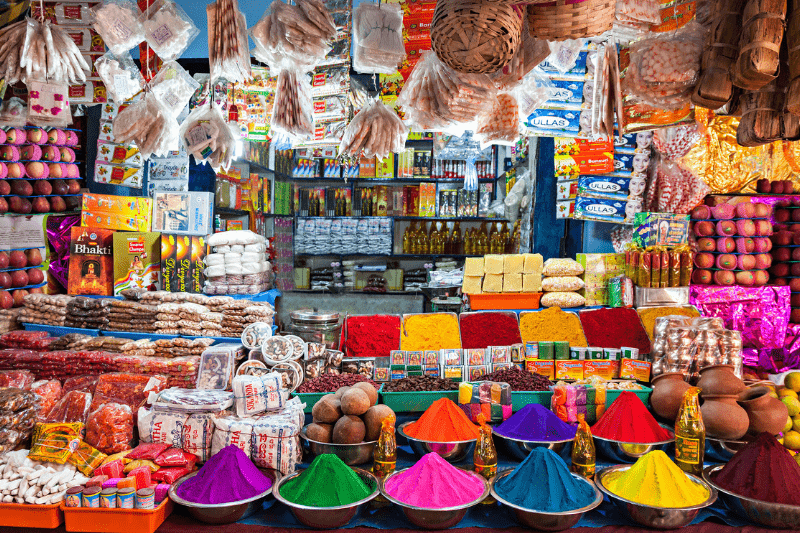 about-page2 Colorful spices and powders in bowls, packaged goods, and assorted market items arranged in a busy, vibrant shop display.