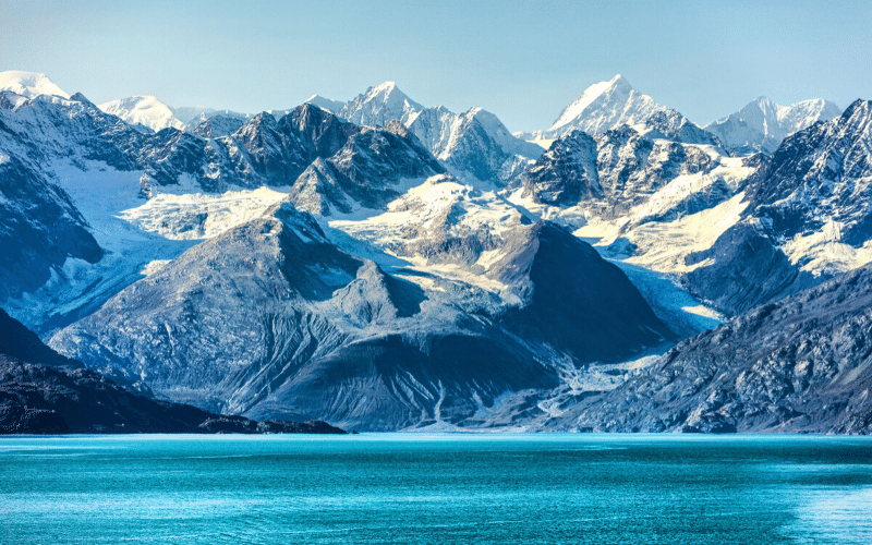 Snow-covered mountains rise behind a blue body of water under a clear sky.