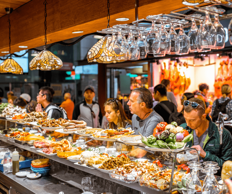 People sit at a crowded market bar eating assorted tapas, with seafood, vegetables, and other dishes visible on the counter under hanging glasses.