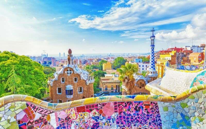 Colorful mosaic benches in Park G&uuml;ell overlooking Barcelona cityscape, with unique buildings, greenery, and a partly cloudy sky in the background.