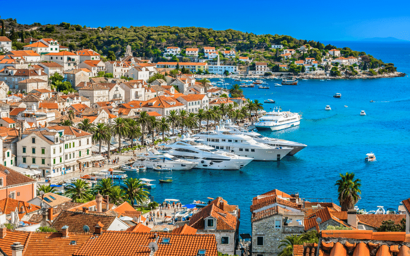 A coastal town with red-roofed buildings, palm trees, and several yachts docked along a blue waterfront under a clear sky.