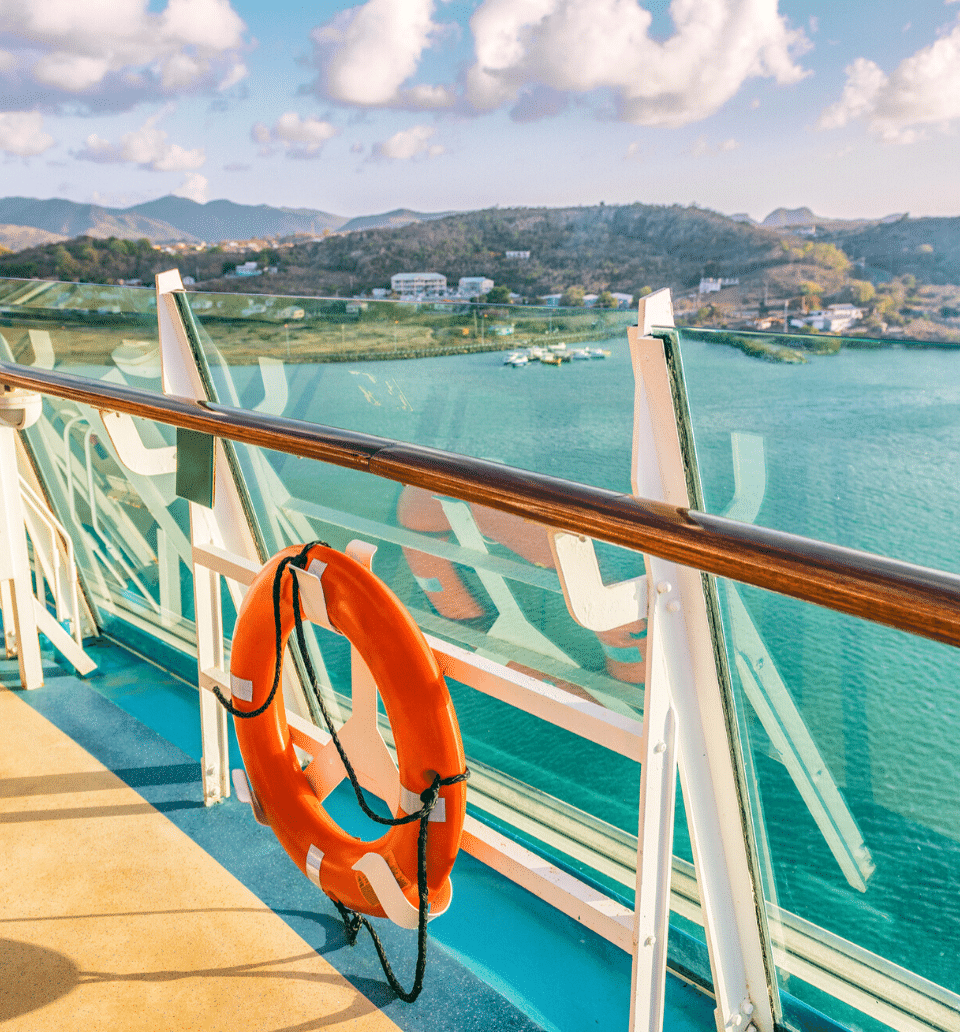 An orange lifebuoy hangs on the railing of a ship deck overlooking calm turquoise water and distant hills under a partly cloudy sky.