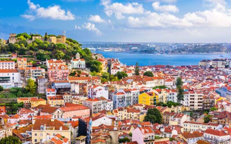 A panoramic view of Lisbon with red-roofed buildings, a hilltop castle, and the Tagus River under a partly cloudy sky.