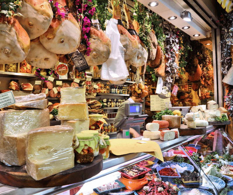 florence-italy A deli counter displays assorted cheeses, cured meats, and jars of preserves, with prosciutto legs hanging above and shelves stocked with food items in the background.