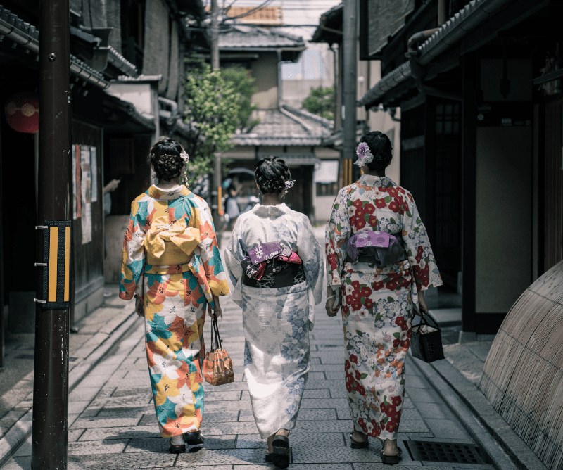Three women wearing colorful kimonos walk down a narrow, traditional Japanese street lined with old buildings.