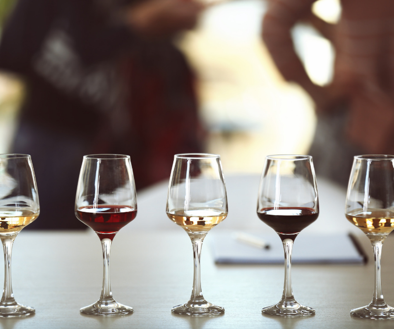 Four wine glasses with varying amounts and colors of wine are arranged in a row on a table, with blurred people in the background.