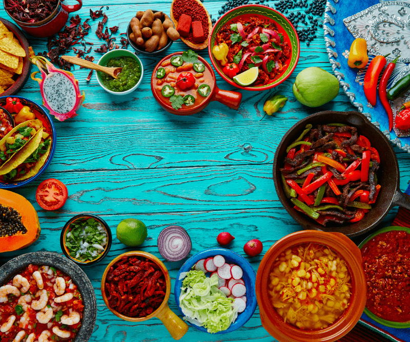 Variety of colorful Mexican dishes, including tacos, soups, saut&eacute;ed vegetables, salsas, and fresh ingredients, arranged on a bright blue wooden table.