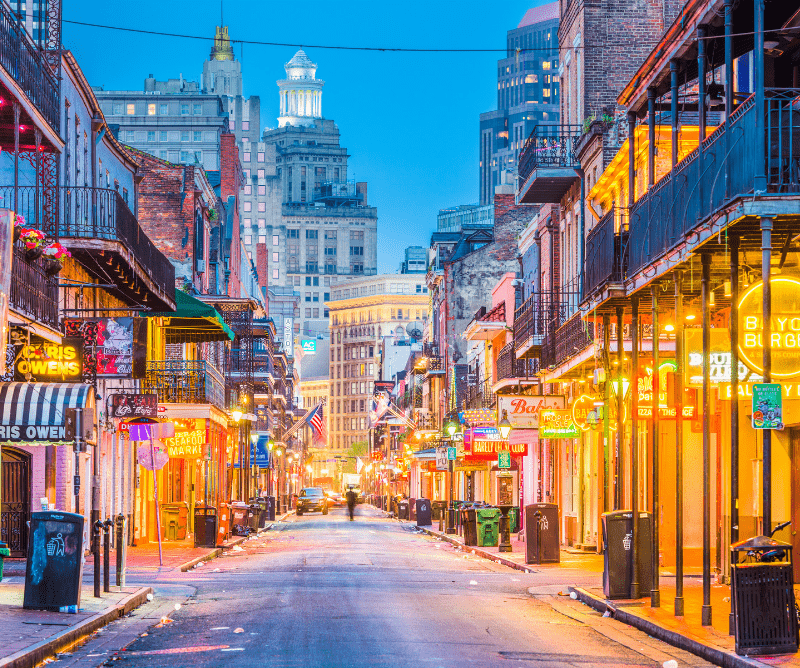 A brightly lit street in New Orleans' French Quarter at dusk, lined with neon signs, historic buildings, and empty except for a distant figure.