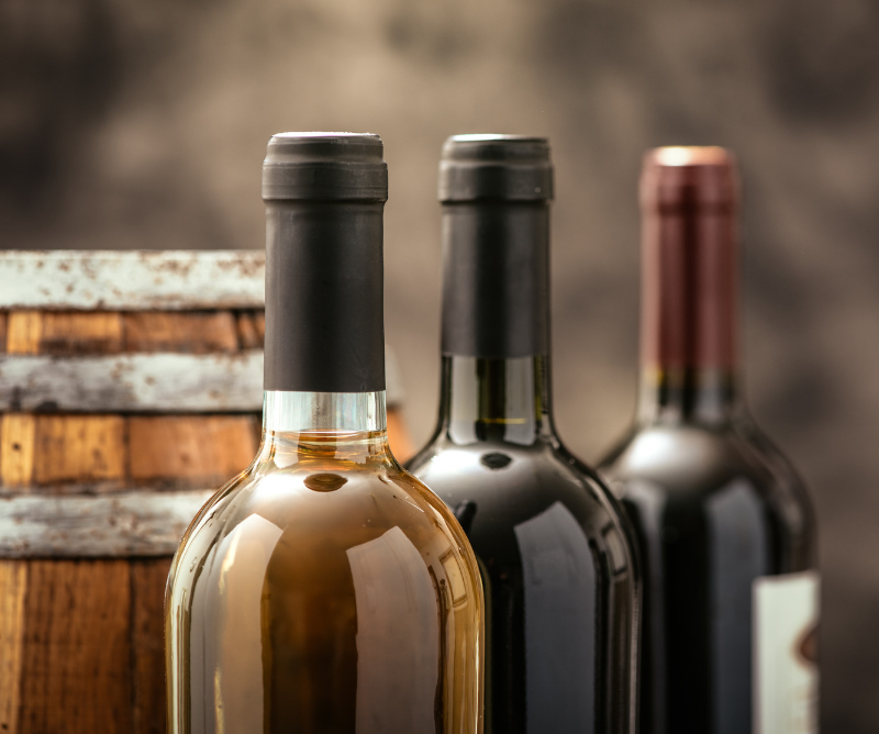 Three bottles of wine, each with a different colored neck foil, are positioned in front of a wooden barrel against a blurred background.