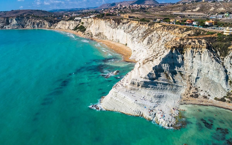 Aerial view of white limestone cliffs and turquoise sea along a coastline, with a sandy beach and scattered buildings on top of the cliffs.