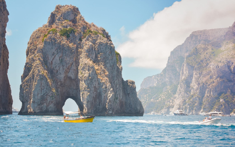 Boats travel on blue water near large rocky formations, one with a natural arch, under a partly cloudy sky.