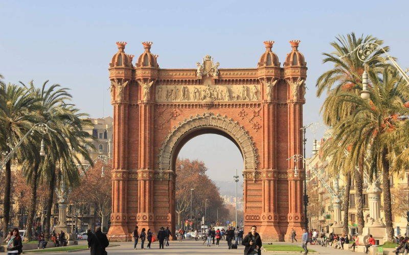 The Arc de Triomf in Barcelona, Spain, is a large red-brick triumphal arch with detailed carvings, situated on a wide pedestrian avenue with people walking and palm trees lining the sides.