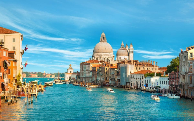 Venice Grand Canal with boats, historic buildings, and the domed Santa Maria della Salute church under a blue sky.