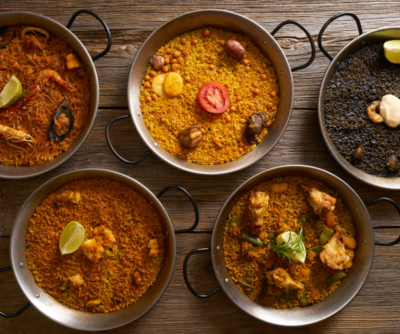 Five pans of assorted Spanish rice dishes, including paella with seafood, chicken, black rice, and various garnishes, arranged on a wooden surface.