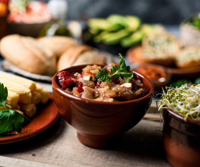 A brown bowl filled with a chunky vegetable dish garnished with mint sits on a table, surrounded by bread, corn, and fresh herbs.