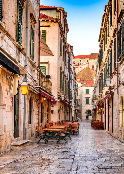 Narrow stone street lined with old buildings, shuttered windows, outdoor tables and chairs, and hanging lamps, under a clear blue sky.