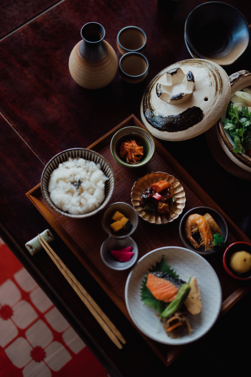 A Japanese meal set on a wooden tray includes rice, assorted side dishes, grilled salmon, pickles, and a teapot, with chopsticks placed to the left.