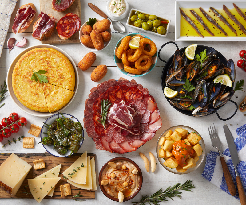An assortment of Spanish tapas on a table, including cured meats, cheese, tortilla, mussels, olives, croquettes, fried rings, anchovies, and various dips.