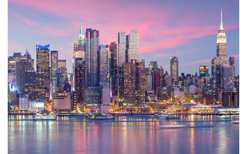 City skyline at dusk with tall buildings illuminated, reflecting on a calm river under a pink and purple sky. City skyline at dusk with tall buildings illuminated, reflecting on a calm river under a pink and purple sky.