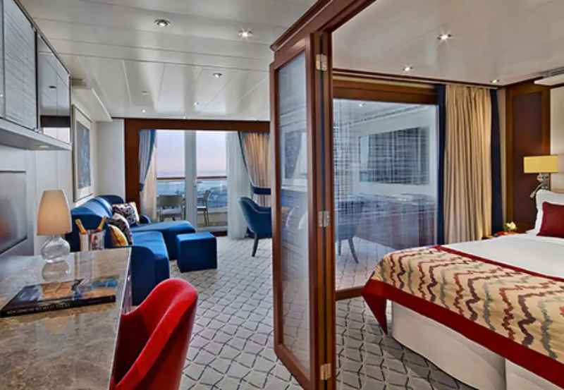 Modern hotel suite with a bedroom, living area, desk, and balcony view of the sea at sunset; glass doors separate the spaces.
