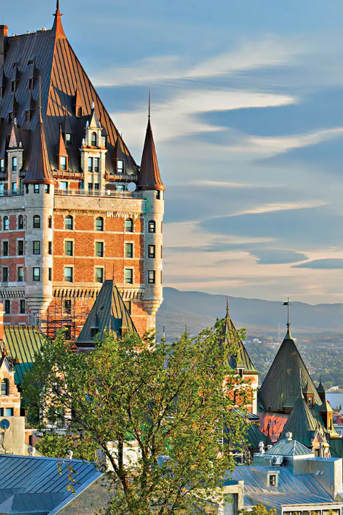 A large historic building with pointed towers and a steep roof stands among trees, with mountains and a river visible in the background under a partly cloudy sky.