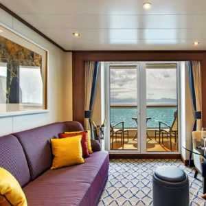 A modern cruise ship cabin with a sofa, colorful cushions, patterned carpet, and a balcony overlooking the ocean with two chairs and a small table.