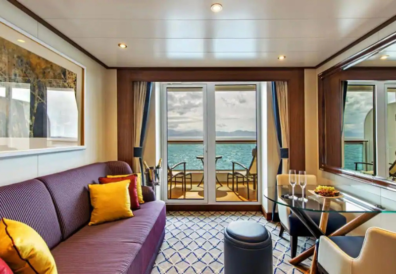 A modern cruise ship cabin with a sofa, pillows, small table set for two, and balcony doors opening to a view of the ocean and distant mountains.