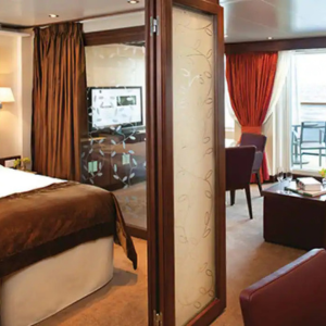A hotel room with a bed, a glass-panel divider, a TV, seating area with chairs and table, floor-to-ceiling windows, and balcony access with red curtains.