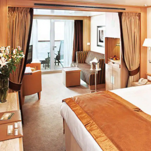 A neatly arranged cruise ship cabin features a bed, sofa, table, armchair, flowers, and a balcony with chairs overlooking the sea.