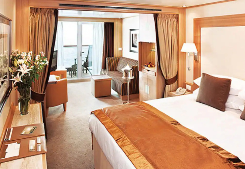 A well-lit hotel room with a double bed, sofa, desk, flowers, and a balcony overlooking the sea. Brown and beige decor dominates the space.