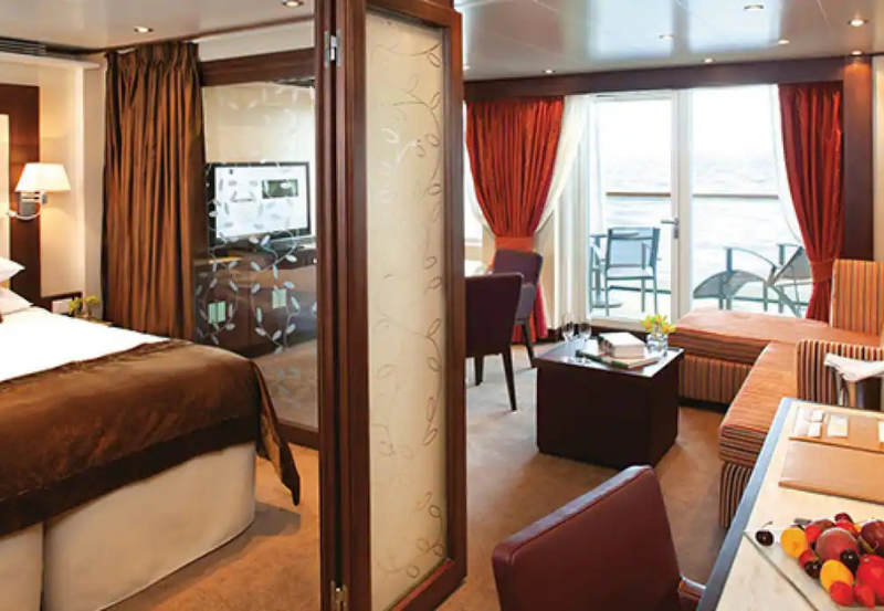 A modern cruise ship cabin features a bed, sitting area, TV, desk, glass door, and balcony with two chairs overlooking the sea.